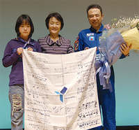 宇宙に持参した間門小児童からの応援フラッグを掲げる古川さん（写真右）と同校児童（左）