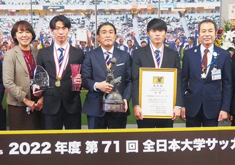 優勝記念パネルの前でトロフィーや賞状を手に笑顔を見せる（左から）森学長、神田選手、安武監督、山内選手、天下谷区長