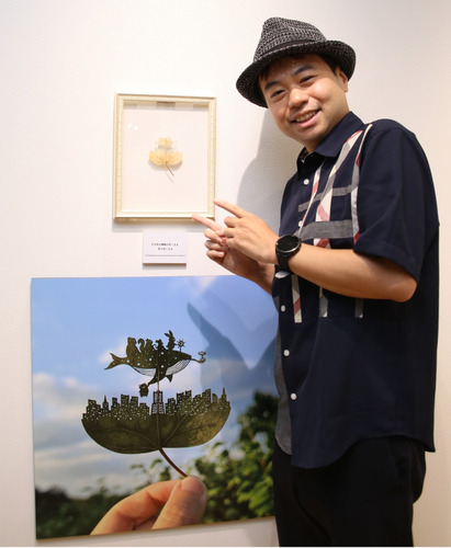 みなとみらいを表現した作品展のメインビジュアルを前に笑顔を見せるリト＠葉っぱ切り絵さん　　【プロフィール】1986年7月14日、神奈川県出身。横浜市保土ケ谷区の富士見台小、岩井原中を経て横浜商科大卒。社会人として働く中、ADHDとの診断を受け、偏った集中力やこだわりを前向きに生かすため、2020年から独学で葉っぱ切り絵の制作活動を開始。SNSに毎日のように投稿する作品が国内外で注目を集め、「情熱大陸」や「徹子の部屋」などテレビでも取り上げられる。現在のSNSフォロワーは約83万人。全国で作品展を開催するほか、2024年には個人美術館「LITO LEAF ART MUSEUM FUKUSHIMA」が福島市にオープン。2025年２月に「やなせたかし文化賞」の大賞を受賞した。