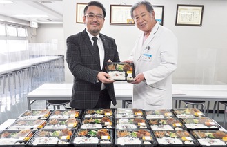 安田代表（写真左）が吉田院長に弁当を手渡した