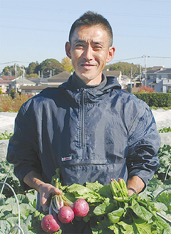 中田中学校裏の畑で野菜を手に吉田さん。栽培から収穫、配達までをほぼ１人で行う