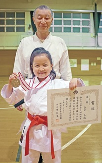 大会の優勝メダルと賞状を持つ益田さん。後ろは志空会の小林志光師範