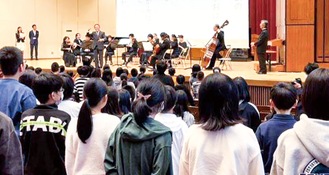 オーケストラの演奏で記念歌を歌う児童