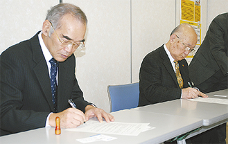 食品提供協定に関する合意書に署名する外狩取締役社長（左）と山崎会長（右）
