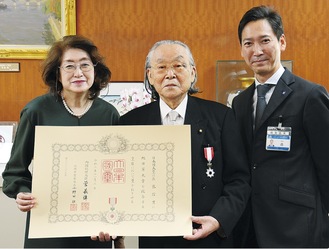勲章を胸に、洋子夫人と勲記を手にする佐藤会長（中央、右＝伝達の代役を務めた森区長）