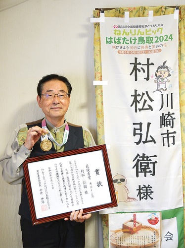 現地でもらった名前入りののぼり旗の前で賞状と金メダルを手にする村松さん