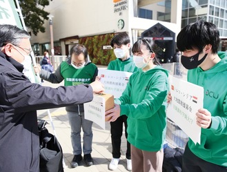 緑の服を着て街頭に立つ学生と、募金した通行人