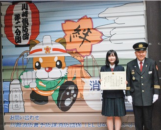 新調したシャッターデザインを背に感謝状を手にする大和さん（左）と小松原団長