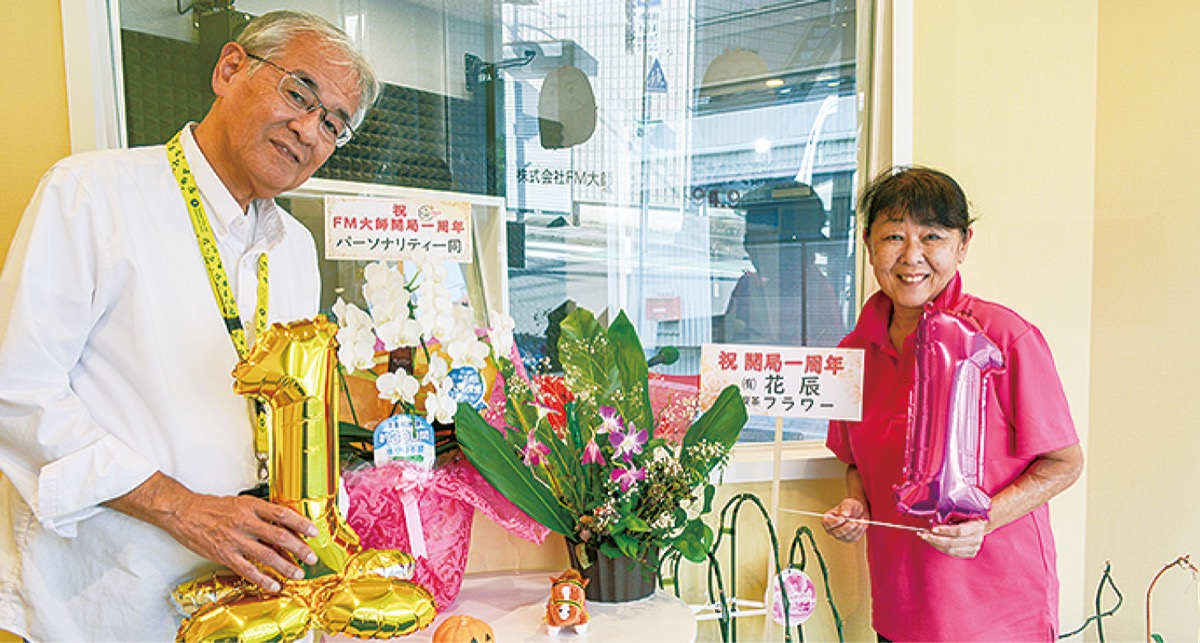 １周年のお祝いの花を前にする清水社長（左）と妻でパーソナリティーの成美さん
