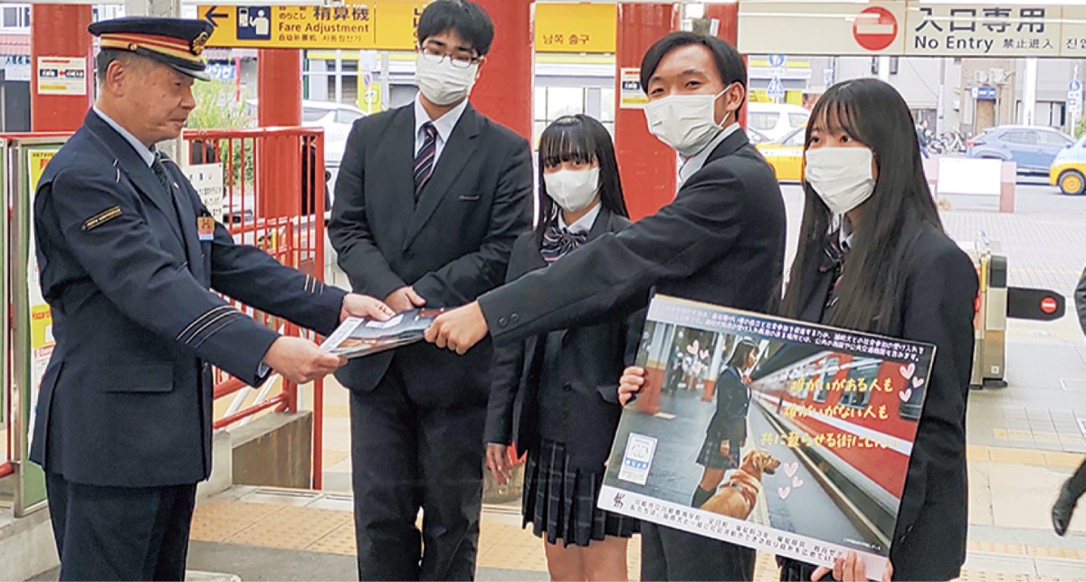 川崎大師駅で鈴木駅長にポスターを渡す様子。右から同高校の斎藤さん、阿部さん、寺崎さん、服部さん。