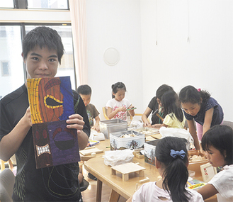 作品を手にする横山さんと作業に集中する児童ら＝8月26日、ウディーショップきこり