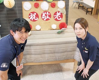 機能訓練士の村上生樹さん（左）と介護職員の玄間由貴さん。「敬老の日」のイベントのために用意したボードの前で