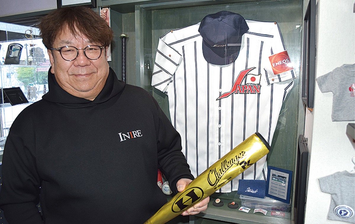 自らデザインしたシンボルマークが入ったユニフォームをバックにバットを握る高橋さん