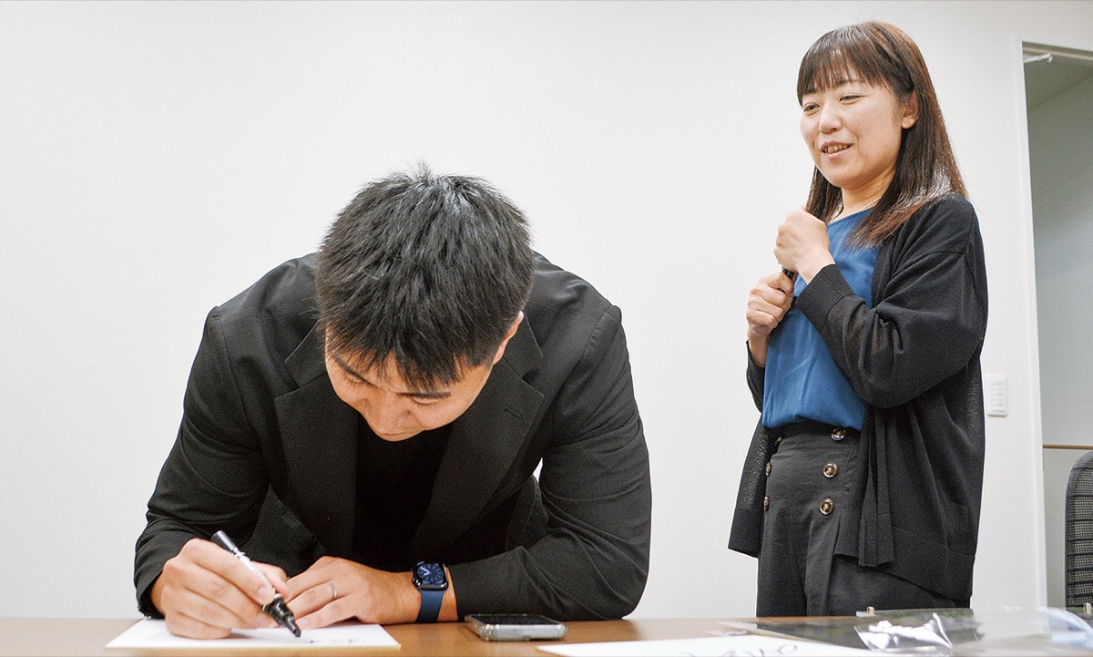▲サインを求める高橋奈己さん（右）はダイナボアーズに熱中している社員の一人。「パートナーとしてのご縁から応援を始めましたが、選手の力強いプレーやスタジアムの熱気に今ではすっかり魅了されています。さらなる躍進を全緑で応援していきます！」