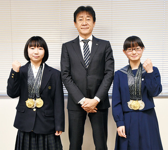 野村教育長を挟んで、3つのメダルを胸にガッツポーズを見せる古川さん（左）と石井さん
