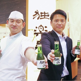 コラボの日本酒を持って独楽寿司八王子オクトーレ店（旭町）の前に立つ高麗さん（左）と西仲さん
