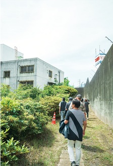 作業療法棟（左手前）の説明を受けながら、高さ約４メートルの塀に沿って歩く参加者