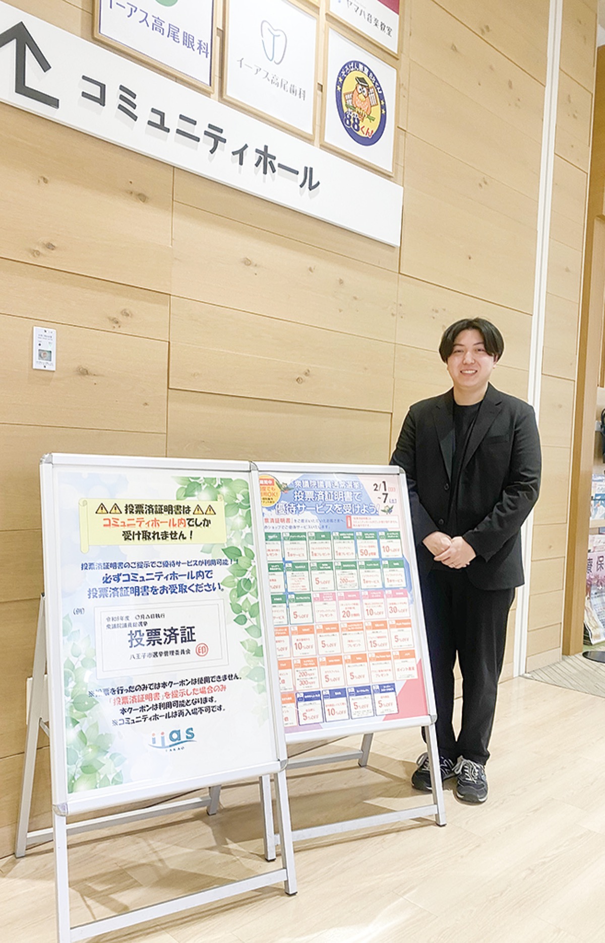 コミュニティホール前に置かれた選挙割ポスターとイーアス高尾の津田さん＝２月３日撮影