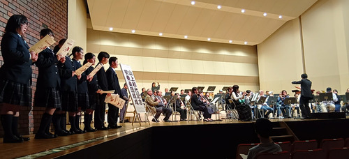 全体合唱で演奏する吹奏楽団（奥）