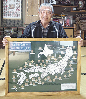 完成作品を手に笑顔の関さん。日本地図に合わせて47都道府県の記念コインを配し、ベニヤ板に嵌め込んだ力作。額縁も手作りだ