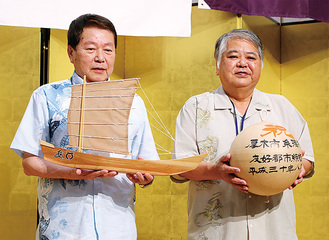 記念品として贈られた「鮎まつり」を象徴する花火玉のレプリカを持つ上原市長（右）と、帆掛け船「サバニ」の模型を手にする小林市長