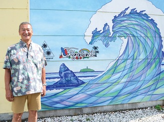店脇の外壁描かれた波をバックに笑顔を見せる藤沢さん＝５月27日、東海岸南のフリュードパワーサーフクラフト