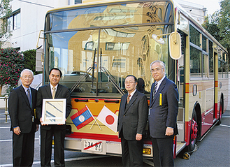 寄贈されるバスの前で、左から同社髙橋幹会長、ラオス大使、瀧野代表、三澤社長