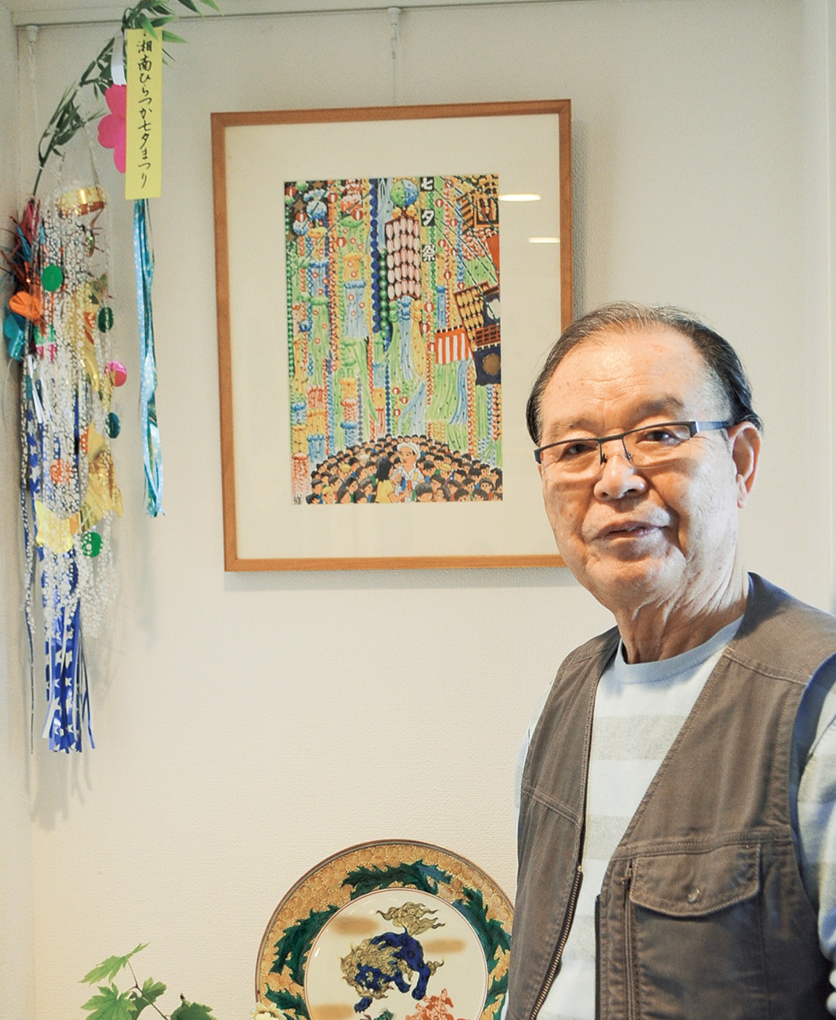 取材を受ける宮代長次さんの息子・臣之さん。後ろには七夕期間中に飾るという絵画と小さな飾りがある