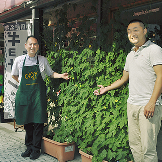 窓を覆う、マツシタ靴店のグリーンカーテン。成長を喜ぶ平井義人さん（左）と松下善彦さん（右）